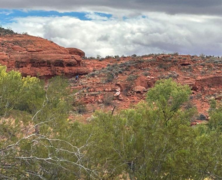 Two mountain bikers on a red rock slope above green brush